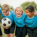 A multi-ethnic group of elementary age children are standing together before their soccer game. One girl is smiling and looking at the camera.